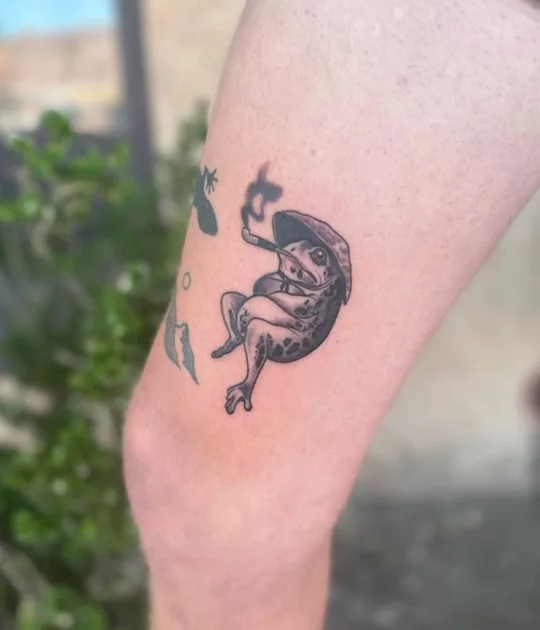 Small black and grey curled figure with floating fragments tattoo on a leg