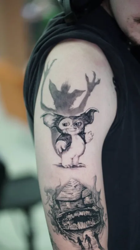 Black and grey large-eared character above a cabin scene tattoo on an upper arm