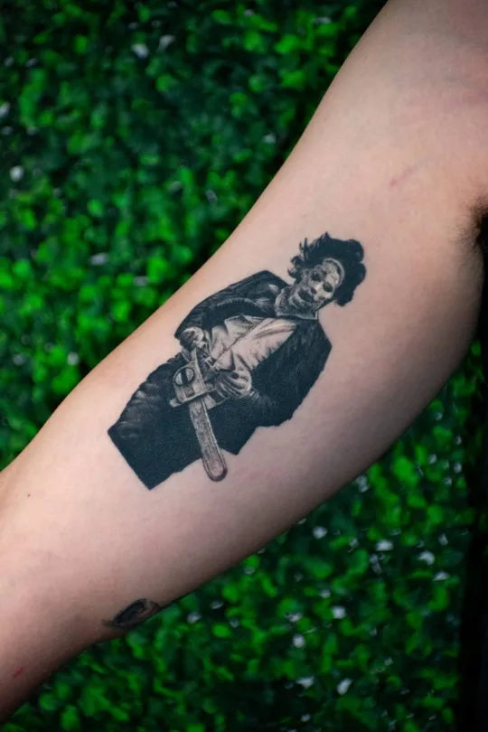 Black and grey portrait of a figure wearing a dark jacket tattoo on an arm